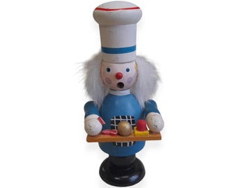 Puede incluir: Figura de chef de madera con gorro blanco y uniforme azul. La figura tiene una expresión de sorpresa y sostiene una bandeja con alimentos. La base es negra y la figura tiene pelo blanco.