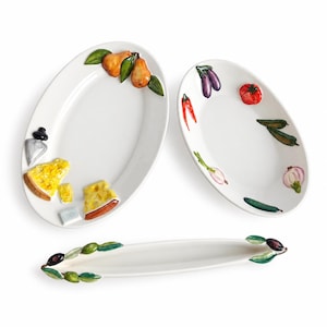 Puede incluir: Tres platos de servicio de cerámica blanca con motivos de alimentos en relieve pintados a mano. Un plato ovalado presenta peras, queso y un cuchillo. Otro plato ovalado muestra berenjena, tomate, pimientos y cebolla. Un plato largo y estrecho está decorado con aceitunas.