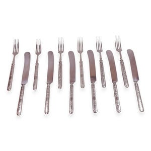 May include: A collection of antique silverware, including forks and knives. The utensils are silver-colored with ornate, engraved handles. The knives have curved blades, and the forks have four tines. The set is arranged on a white background.