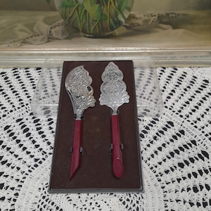 Peut inclure: Deux couteaux à beurre en argent avec des motifs ornés et des manches rouges dans une boîte bordeaux.