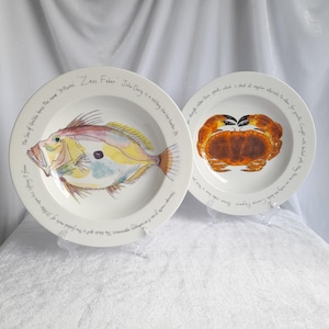 May include: Two white ceramic plates with illustrations of marine life. One plate features a colorful fish, the other a crab. Text encircles the rims of both plates. The plates are displayed on clear stands.