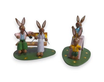 Peut inclure: Figurines de lapins de Pâques en bois. Une scène montre deux lapins sur un banc, l'un fumant une pipe. L'autre scène montre trois lapins sur un toboggan. Les figurines sont peintes de différentes couleurs.