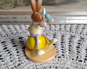 May include: A wooden bunny rabbit figurine holding a paintbrush and sitting on a yellow and purple Easter egg. The bunny is painted in shades of brown, yellow, and white. The figurine is sitting on a wooden base.