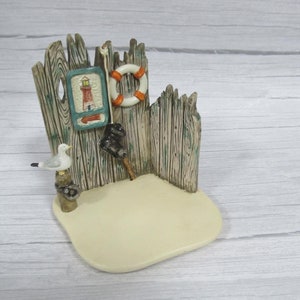 May include: A miniature wooden fence with a lighthouse sign, a life preserver, and a seagull perched on a post. The fence is painted green and brown, and the base is a light beige color.