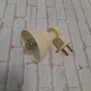 Puede incluir: Una lámpara de pared de plástico blanco con una pantalla blanca y una bombilla transparente. La lámpara está diseñada para montarse en una pared.