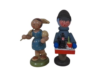May include: Two wooden figurines. One is a girl with a blue dress with white polka dots, holding a stick. The other is a man in a blue coat and black hat, holding a tray with a small black object and a red cup.