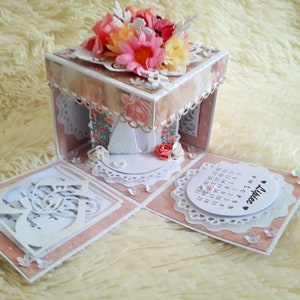 Può includere: Una scatola di carta rosa e bianca con un design floreale e un bordo di pizzo bianco. La scatola è aperta e rivela una figura di carta bianca di una sposa e uno sposo. La scatola ha anche un calendario con la parola "Alice" scritta su di essa.