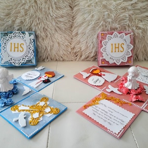 Può includere: Due scatole esplosive aperte decorate per una celebrazione della Prima Comunione. La scatola blu presenta una statuetta di angelo bianco, una croce dorata e colombe bianche. La scatola rosa presenta una statuetta di angelo bianco, un calice dorato e colombe bianche. Entrambe le scatole hanno un cerchio di pizzo bianco con le lettere "IHS" in oro.