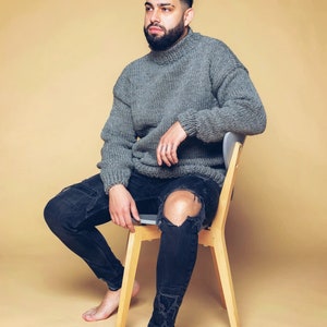 Puede incluir: Una persona con un jersey de cuello alto gris de punto grueso y vaqueros negros desgastados. La persona est&aacute; sentada en una silla de madera sobre un fondo beige. Los vaqueros tienen rodillas rasgadas.