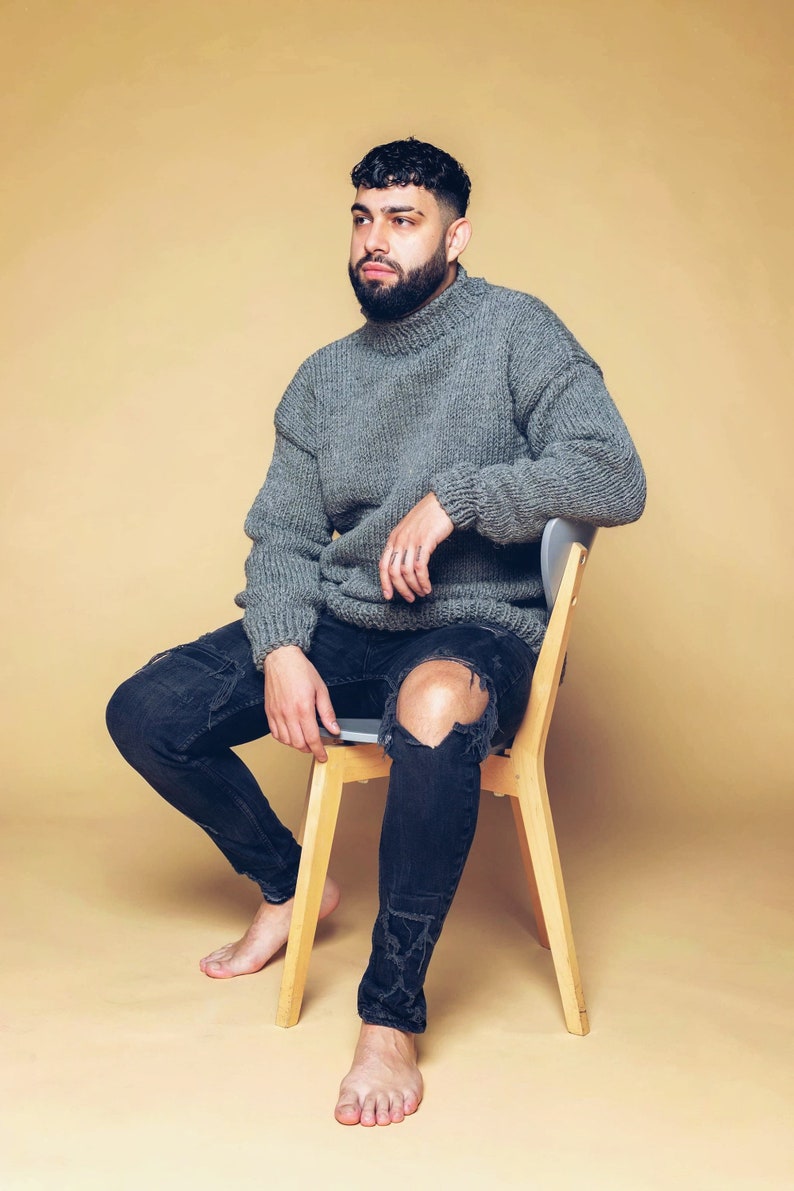 Puede incluir: Una persona con un jersey de cuello alto gris de punto grueso y vaqueros negros desgastados. La persona est&aacute; sentada en una silla de madera sobre un fondo beige. Los vaqueros tienen rodillas rasgadas.