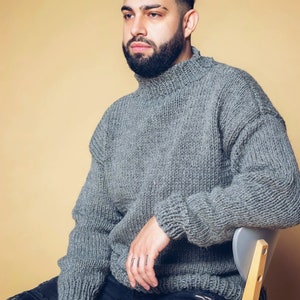 Puede incluir: Un su&eacute;ter gris de cuello alto de gran tama&ntilde;o con una textura de punto grueso. El su&eacute;ter se combina con unos vaqueros negros desgastados. La persona est&aacute; sentada en una silla gris sobre un fondo de color beige claro.