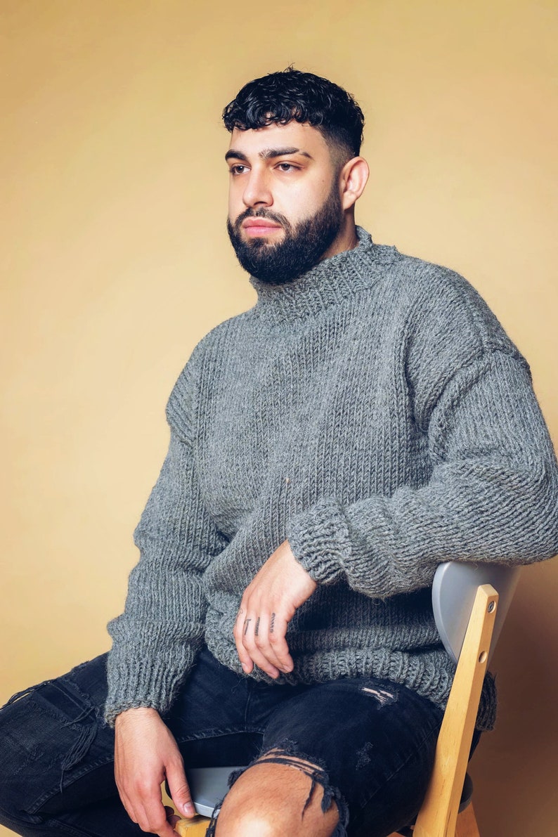 Puede incluir: Un su&eacute;ter gris de cuello alto de gran tama&ntilde;o con una textura de punto grueso. El su&eacute;ter se combina con unos vaqueros negros desgastados. La persona est&aacute; sentada en una silla gris sobre un fondo de color beige claro.