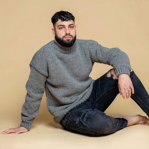 Puede incluir: Un jersey gris de cuello alto con una textura de punto grueso. El jersey se combina con unos vaqueros negros desgastados. La persona est&aacute; sentada sobre un fondo neutro.