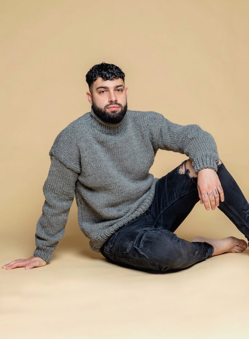 Puede incluir: Un jersey gris de cuello alto con una textura de punto grueso. El jersey se combina con unos vaqueros negros desgastados. La persona est&aacute; sentada sobre un fondo neutro.