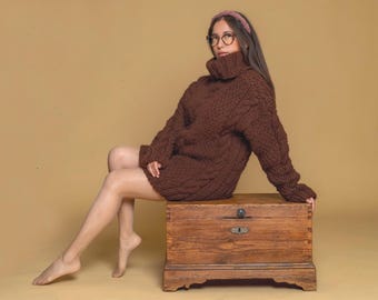 May include: A dark brown, chunky knit sweater featuring a prominent cable knit pattern, a high turtleneck collar, and long sleeves. The garment is worn over a wooden chest with a metal latch and keyhole, showcasing a textured, thick yarn construction.