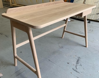 Op de afbeelding: Een licht houten bureau met een eenvoudig ontwerp. Het bureau heeft een rechthoekig blad met een kleine lade aan de rechterkant. De poten zijn schuin en hebben een licht gebogen vorm.