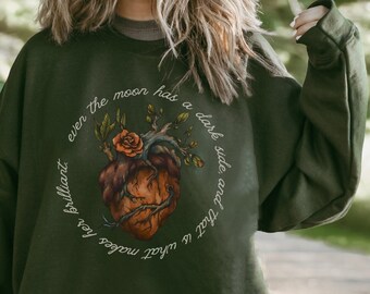 Op de afbeelding: Een donkergroen sweatshirt met een grafische print van een hart met wijnstokken en een roos. De tekst rond het hart luidt "Zelfs de maan heeft een donkere kant en dat is wat haar briljant maakt."