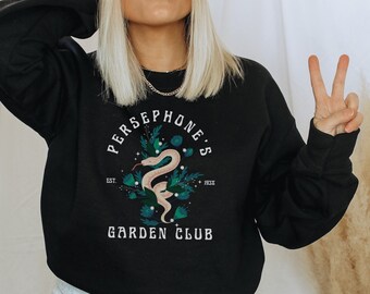 Peut inclure: Sweat-shirt noir avec un imprimé graphique blanc d'un serpent dans un jardin avec le texte "Persephone's Garden Club"