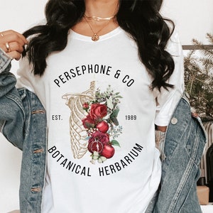 May include: White t-shirt with a floral and skeletal design. The text reads "PERSEPHONE & CO EST. 1989 BOTANICAL HERBARIUM".