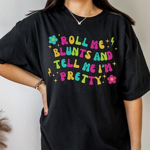May include: Black t-shirt with the text "Roll Me Blunts and Tell Me I'm Pretty" in colourful letters with flowers and lightning bolts.