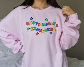 May include: Pink oversized sweatshirt with the text "Emotionally Exhausted" in colorful letters with flower accents.