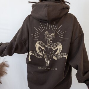 May include: Brown hooded sweatshirt with a graphic print of a skull with snakes and flowers. The text "Memento Mori Vixi" is printed below the graphic.