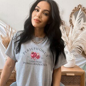 May include: A gray t-shirt with a graphic of a pomegranate and the text "PERSEPHONE'S EST. 1963 FARM FRESH POMEGRANATES".