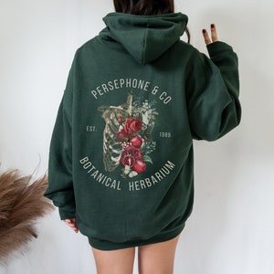 May include: A dark green hooded sweatshirt with a floral and skeletal design on the back. The design features a ribcage with flowers and fruit, and the text "PERSEPHONE & CO EST. 1989 BOTANICAL HERBARIUM".