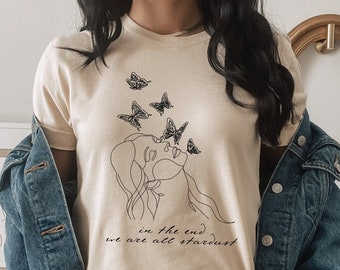 May include: A beige t-shirt with a black line drawing of a woman's face with butterflies around her. The text "in the end we are all stardust" is printed below the drawing.
