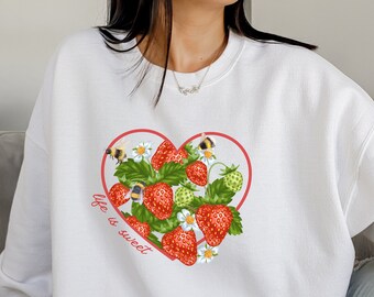 Könnte beinhalten: Weißer Pullover mit einem roten Herz-Motiv mit Erdbeeren, Bienen und grünen Blättern. Der Text "Life is sweet" ist in einer kursivschriftlichen Schrift unter dem Herzen geschrieben.