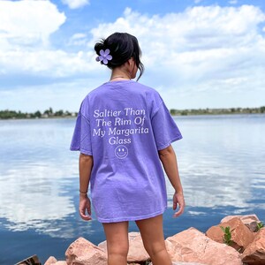 Może przedstawiać: Kobieta w fioletowym t-shircie z napisem "Saltier Than The Rim Of My Margarita Glass" i uśmiechniętą buźką. Stoi przed zbiornikiem wodnym z błękitnym niebem i białymi chmurami w tle.