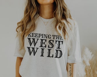 Puede incluir: Una camiseta blanca de gran tamaño con texto negro que dice "KEEPING THE WEST WILD". La modelo lleva pantalones cortos de ciclista grises.