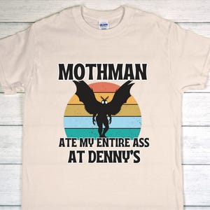 以下が含まれることがあります： クリーム色の半袖Tシャツ。レトロな夕日の円を背景に、黒いモスマンのシルエットが中央に描かれています。グラフィックの上下には、太い黒文字で「MOTHMAN ATE MY ENTIRE ASS AT DENNY'S」というテキストがプリントされています。