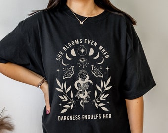 May include: Black oversized t-shirt with a graphic design featuring a rose, snake, and moon phases. The text reads "She blooms even when darkness engulfs her."