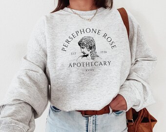Peut inclure: Un sweat-shirt gris avec un imprimé graphique noir d'un visage de femme dans un cercle. Le texte "PERSEPHONE ROSE EST 1936 APOTHECARY XLVII" est imprimé autour du cercle.