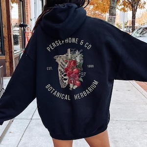 May include: Black hooded sweatshirt with a graphic print of a skeleton with flowers and the text "Persephone & Co. Est. 1999 Botanical Herbarium".