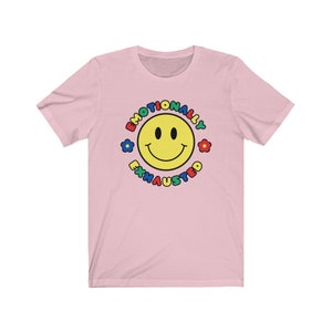 May include: Pink short-sleeved t-shirt with a yellow smiley face in a circle. The circle has the text "Emotionally Exhausted" around the smiley face in rainbow colors.