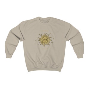 Peut inclure: Un sweat-shirt beige clair avec un motif soleil et &eacute;toiles dor&eacute;s. Le soleil a un visage avec deux yeux et une bouche. Le texte "Tout ira bien" est &eacute;crit sous le soleil.