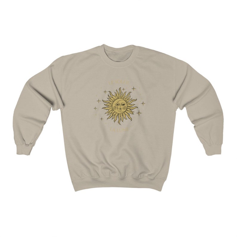 Peut inclure: Un sweat-shirt beige clair avec un motif soleil et &eacute;toiles dor&eacute;s. Le soleil a un visage avec deux yeux et une bouche. Le texte "Tout ira bien" est &eacute;crit sous le soleil.