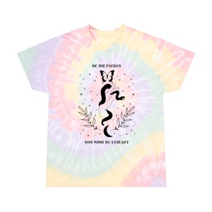 以下が含まれることがあります： パステルカラーのタイダイTシャツ。シャツには、頭の上に蝶が乗った蛇を手に持った手のグラフィックデザインが施されています。グラフィックの下には「BE THE ENERGY YOU WISH TO ATTRACT」というテキストが印刷されています。