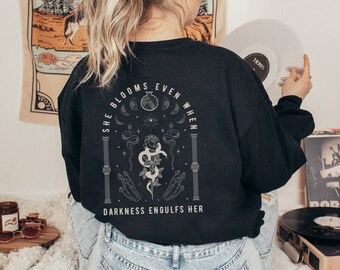 Peut inclure: Sweat-shirt noir avec un imprimé graphique blanc d'une rose, d'un serpent et de phases lunaires. Le texte sur le sweat-shirt est "She blooms even when darkness engulfs her."