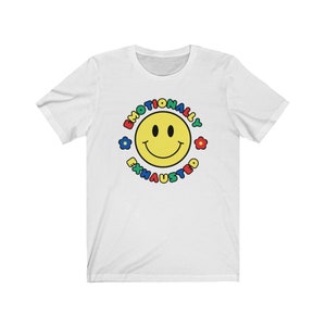 May include: White t-shirt with a yellow smiley face surrounded by the text "Emotionally Exhausted" in a colorful circle.