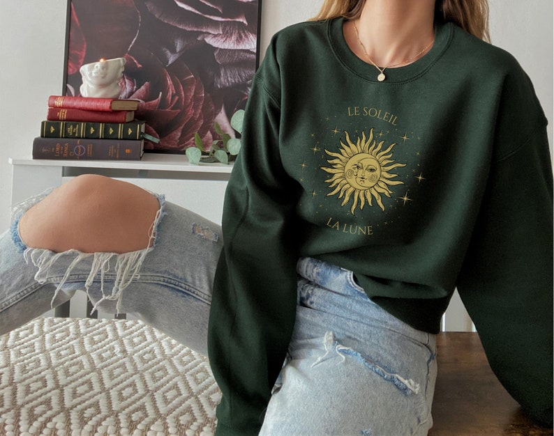 Peut inclure: Un sweat-shirt vert fonc&eacute; avec un motif de soleil et de lune avec le texte "LE SOLEIL" et "LA LUNE" en lettres dor&eacute;es. Le mod&egrave;le porte un jean bleu.