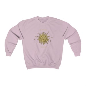 Peut inclure: Un sweat-shirt rose p&acirc;le avec un motif soleil et &eacute;toiles dor&eacute;s. Le soleil a un visage et le texte "Be Soleil La Lune" est imprim&eacute; sous le soleil.