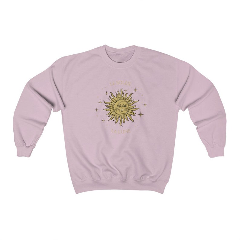 Peut inclure: Un sweat-shirt rose p&acirc;le avec un motif soleil et &eacute;toiles dor&eacute;s. Le soleil a un visage et le texte "Be Soleil La Lune" est imprim&eacute; sous le soleil.