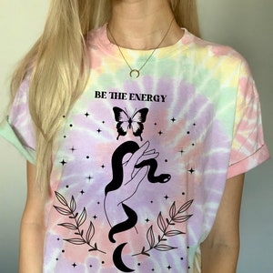 以下が含まれることがあります： パステルカラーのタイダイTシャツ。シャツには、蛇を握った手、蝶、そして「BE THE ENERGY YOU WISH TO ATTRACT」というテキストの白黒のグラフィックが描かれています。