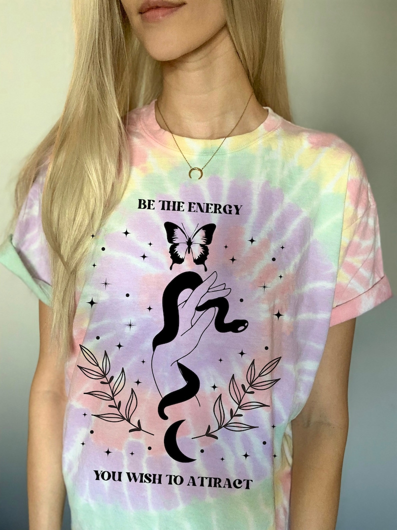 以下が含まれることがあります： パステルカラーのタイダイTシャツ。シャツには、蛇を握った手、蝶、そして「BE THE ENERGY YOU WISH TO ATTRACT」というテキストの白黒のグラフィックが描かれています。