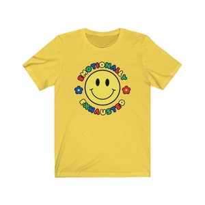May include: Yellow short-sleeved t-shirt with a colorful smiley face graphic and the text "Emotionally Exhausted" around the face.