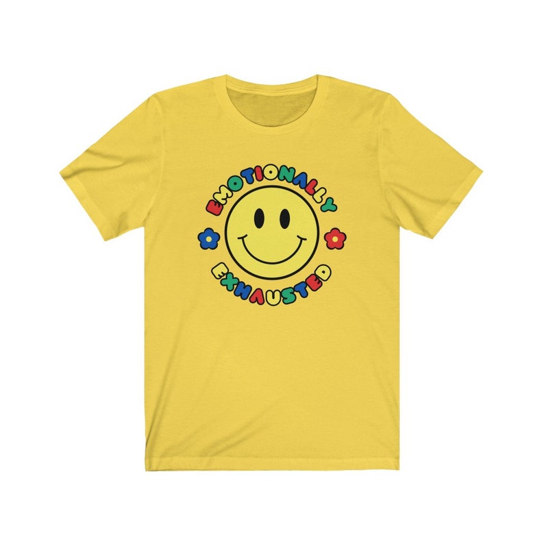 May include: Yellow short-sleeved t-shirt with a colorful smiley face graphic and the text "Emotionally Exhausted" around the face.