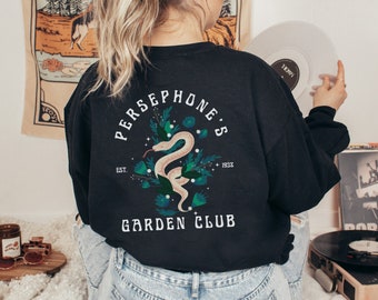 Peut inclure: T-shirt à manches longues noire avec un imprimé graphique d'un serpent blanc avec des feuilles et des fleurs vertes. Le texte "PERSEPHONE'S GARDEN CLUB" est imprimé sur le t-shirt. Le texte "EST. 1938" est également imprimé sur le t-shirt.